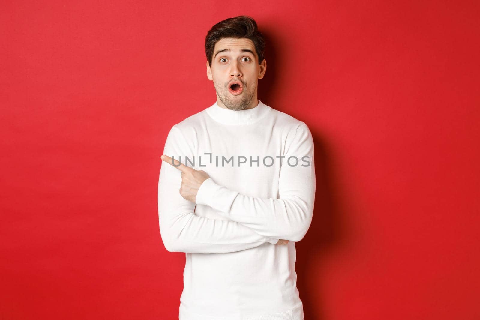 Concept of winter holidays, christmas and lifestyle. Surprised handsome guy pointing finger at new year advertisement, pointing finger left and drop jaw amazed, standing over red background.