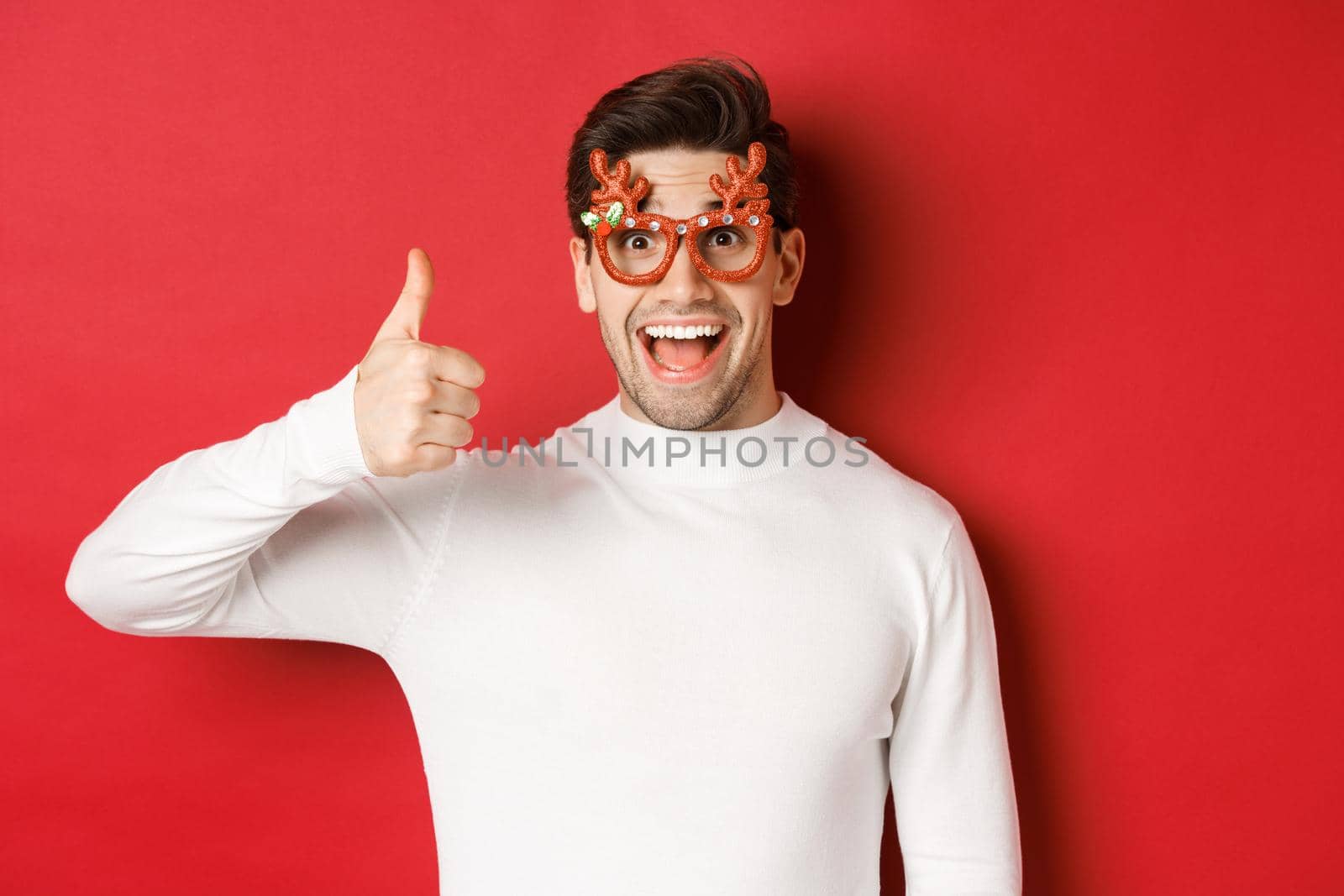 Close-up of handsome young man in party glasses and white sweater, showing thumb-up in approval, enjoying new year holidays, standing over red background.