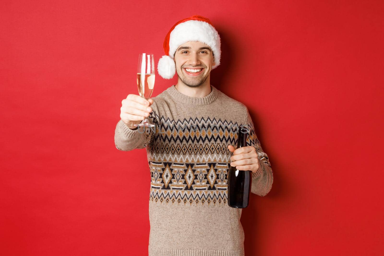 Concept of winter holidays, new year and celebration. Portrait of handsome man in santa hat and sweater, holding champagne, raising glass and saying cheers on christmas party.