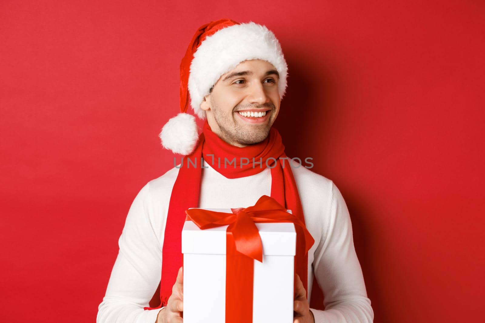 Concept of winter holidays, christmas and lifestyle. Close-up of attractive man in santa hat and scarf, holding new year gift, looking right and smiling, standing over red background.