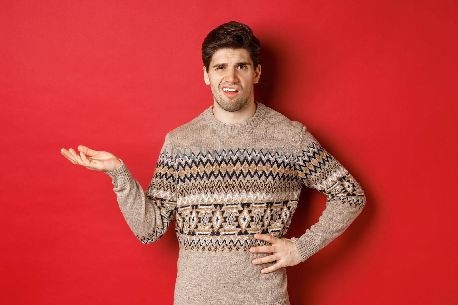 Image of confused and disappointed handsome man, complaining about christmas party, raising hand and looking bothered, standing over red background.