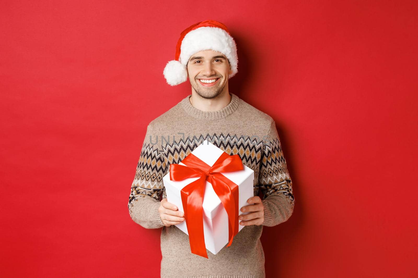 Image of handsome bearded man in winter sweater and santa hat, holding christmas gift and smiling, have a present wrapped with ribbon, standing over red background.