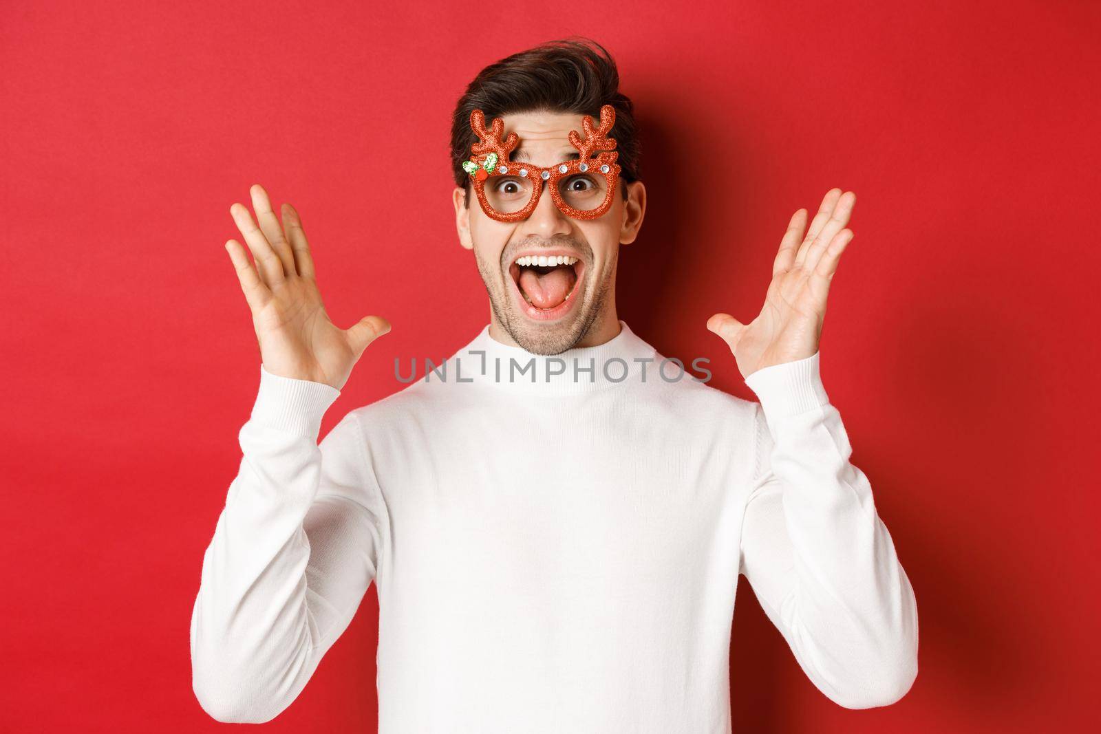Close-up of surprised handsome guy in white sweater and party glasses, hear amazing christmas promo offer, rejoicing and standing over red background.