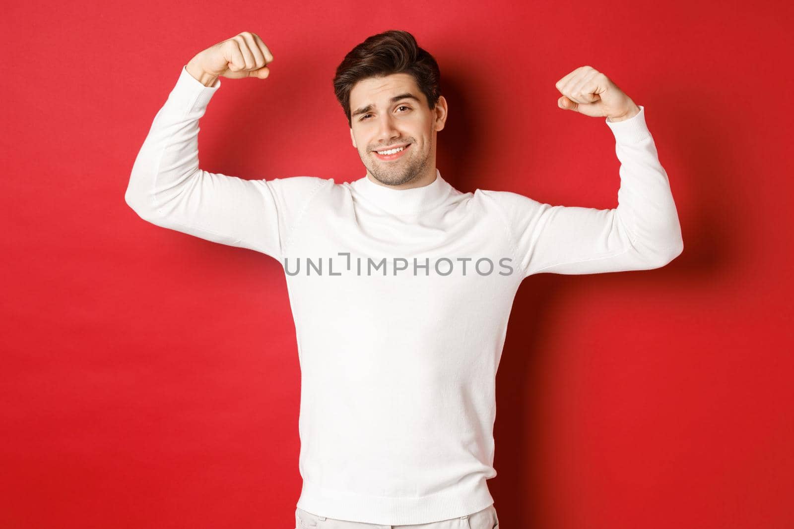 Portrait of smiling handsome man in white sweater, flexing biceps and bragging with strength, show-off strong muscles after workout, standing over red background.