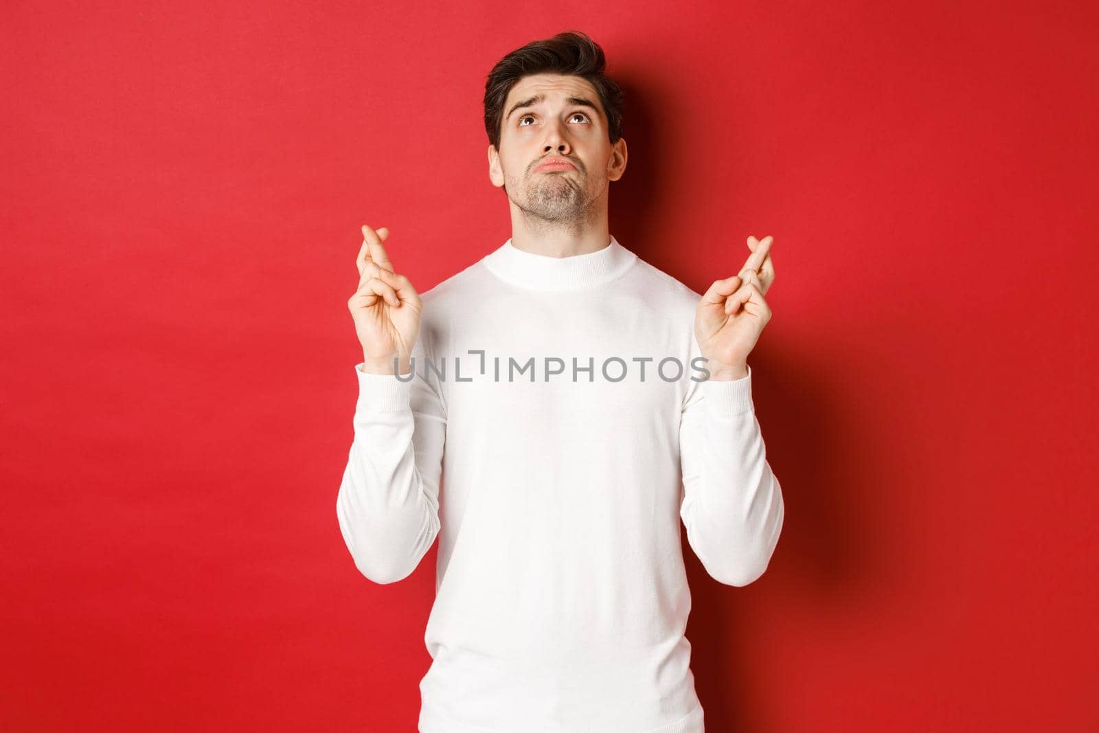 Portrait of sad and hopeful man making a wish, begging god and looking up, crossing fingers for good luck, standing over red background.