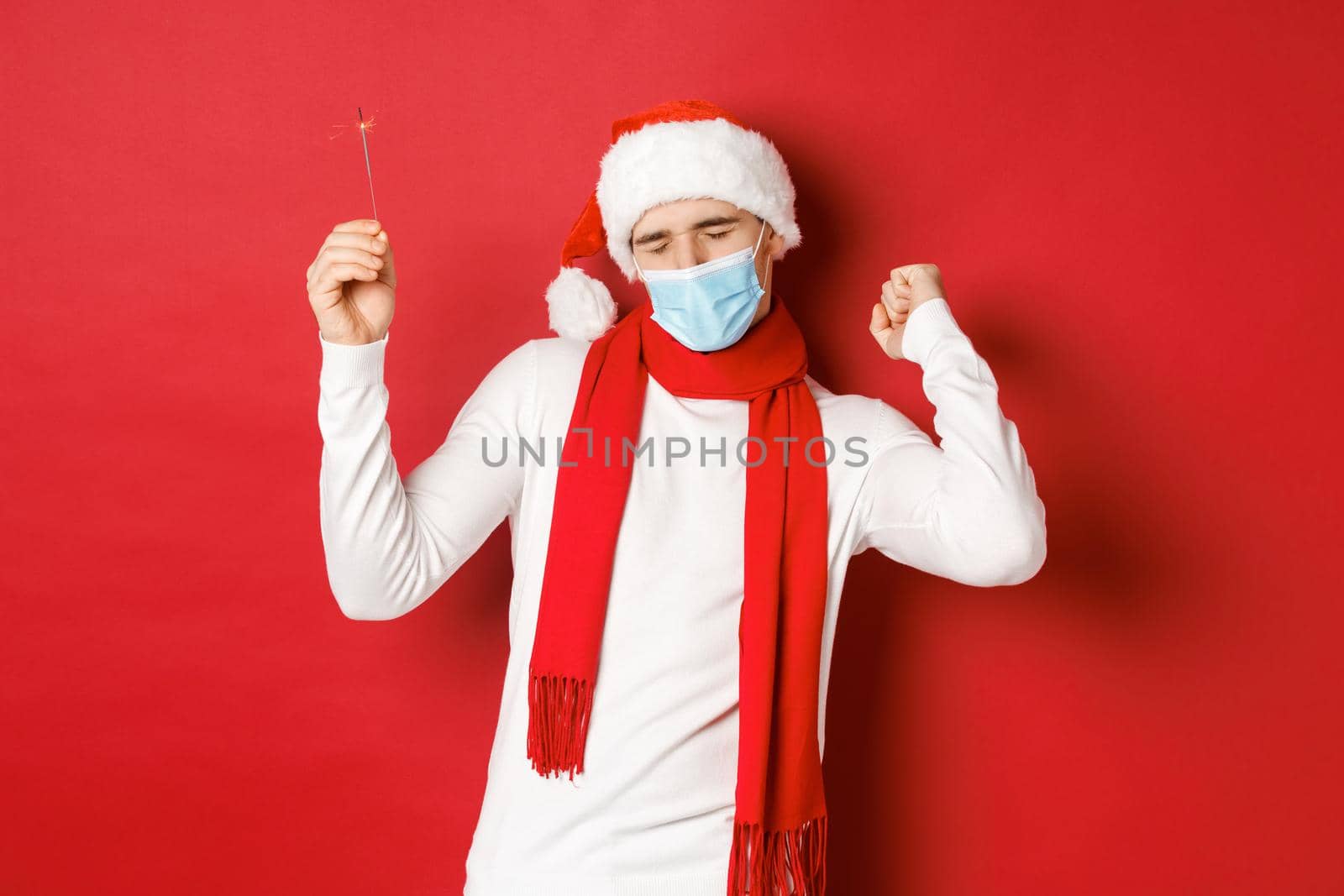 Concept of covid-19, christmas and holidays during pandemic. Happy man celebrating new year at party, wearing medical mask and santa hat, dancing with sparkler against red background.
