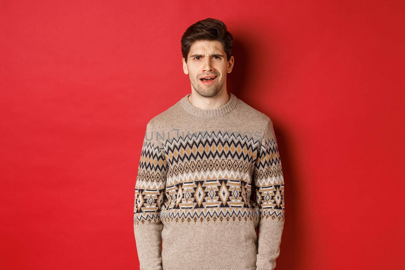 Portrait of confused adult man in christmas sweater, standing disappointed during new year holidays, dont understand something, standing over red background by Benzoix