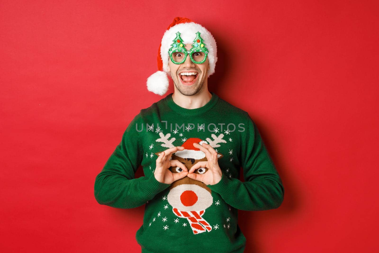 Portrait of carefree handsome man in santa hat and party glasses, making fun of his christmas sweater, looking happy over red background.