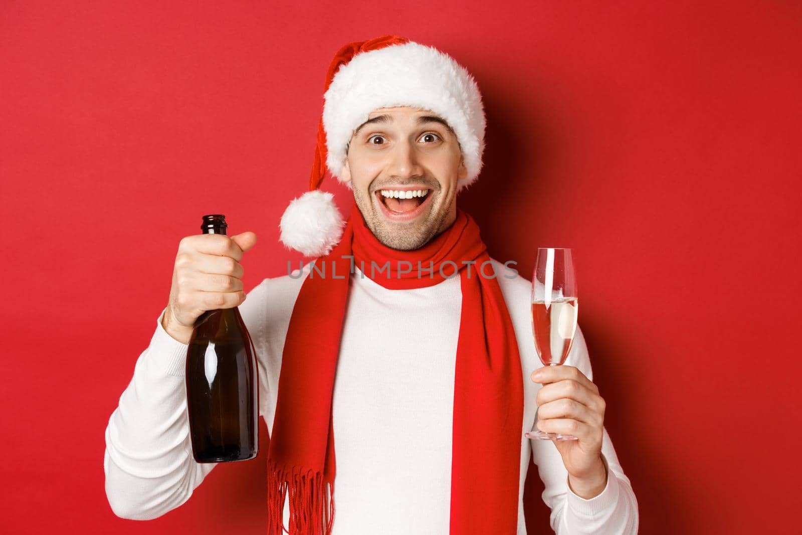 Concept of winter holidays, christmas and lifestyle. Close-up of cheerful handsome man, holding champagne bottle and glass, celebrating new year, standing over red background.