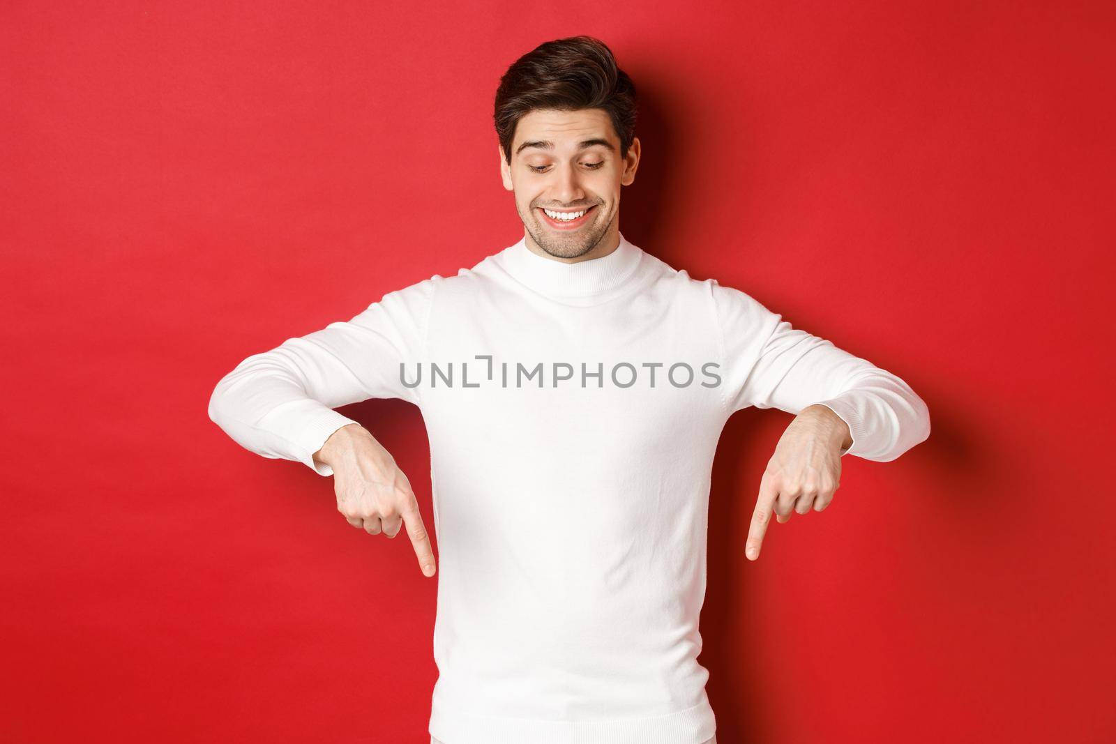 Image of dreamy good-looking man in white sweater, looking and pointing fingers down at copy space, advertisement about new year and holidays, standing over red background.