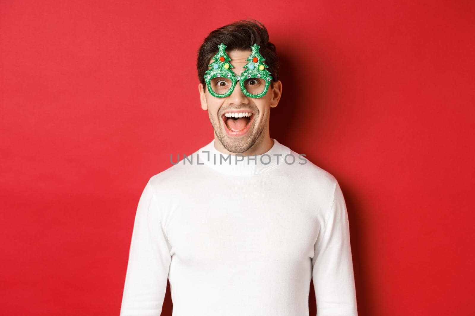 Concept of christmas, winter holidays and celebration. Close-up of handsome young man enjoying new year, wearing party glasses and looking amazed at camera, standing over red background.