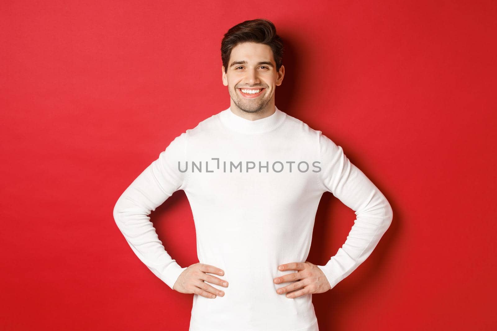 Portrait of handsome man with bristle, wearing white sweater, smiling and looking confident, standing against red background. Concept of new year and winter holidays