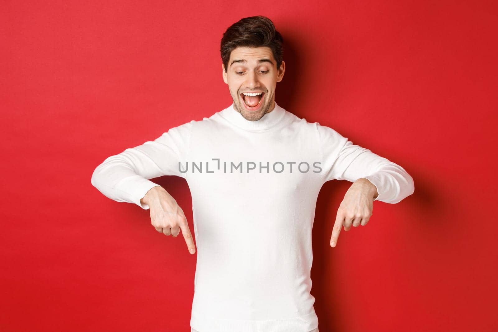 Portrait of happy good-looking man, pointing fingers down and looking at advertisement with excited smile, standing over red background by Benzoix