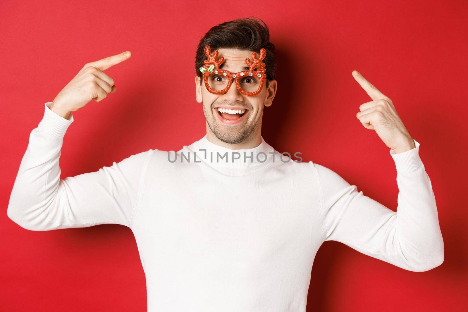 Handsome happy guy in white sweater, pointing at his christmas party glasses, celebrating new year and having fun, standing over red background.