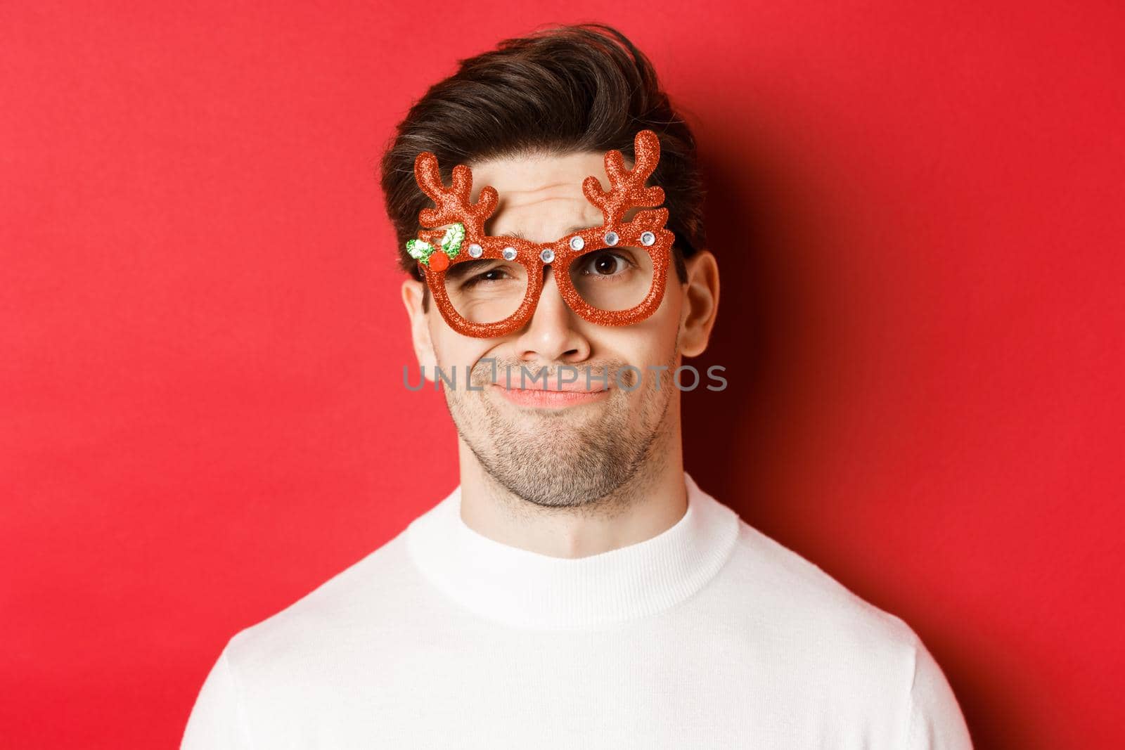 Concept of winter holidays, christmas and celebration. Close-up of skeptical handsome man in party glasses, looking doubtful and unamused, standing against red background.