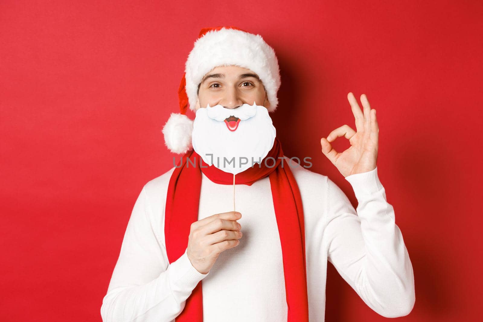 Concept of christmas, winter holidays and celebration. Pleased handsome man in santa hat, holding long white beard mask and showing okay sign, standing over red background by Benzoix