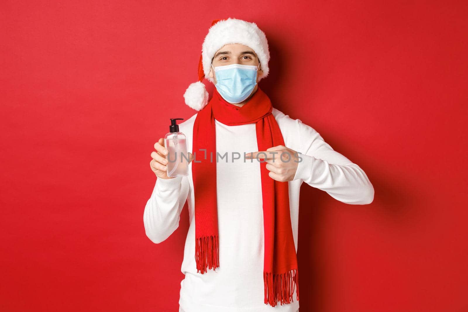 Concept of covid-19, christmas and holidays during pandemic. Handsome happy man in santa hat and medical mask, recommending use hand sanitizer, standing over red background by Benzoix