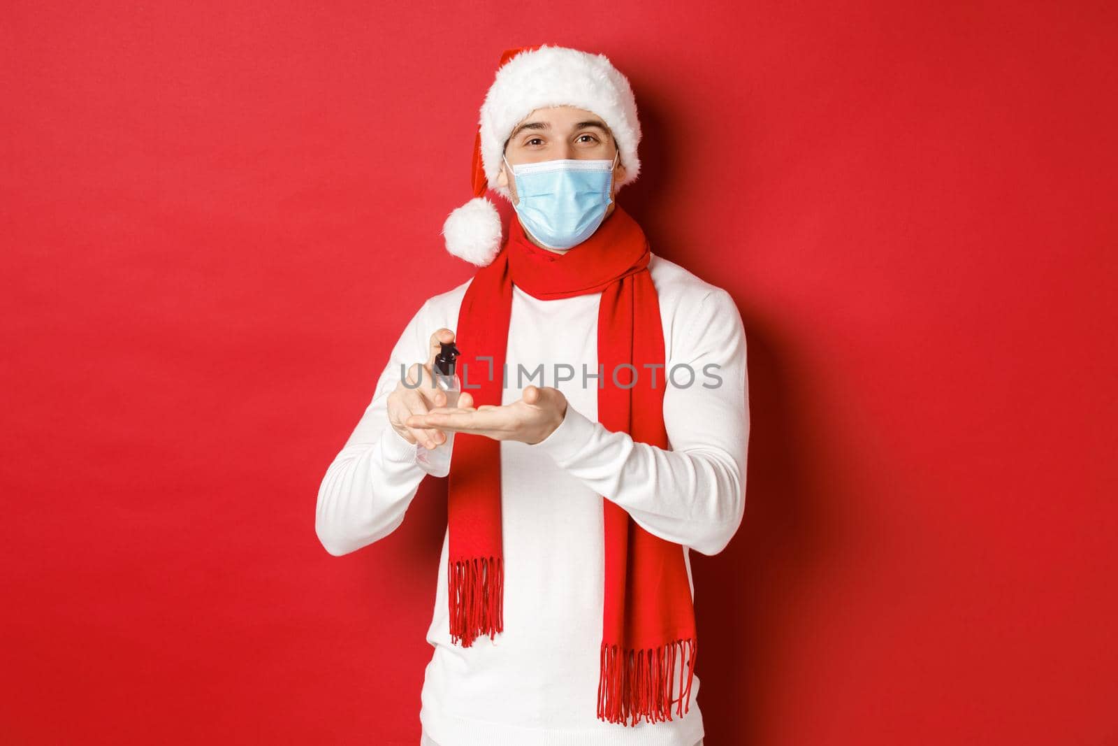 Concept of covid-19, christmas and holidays during pandemic. Happy young man in santa hat and medical mask, sanitize hands with antiseptic and smiling, standing over red background.
