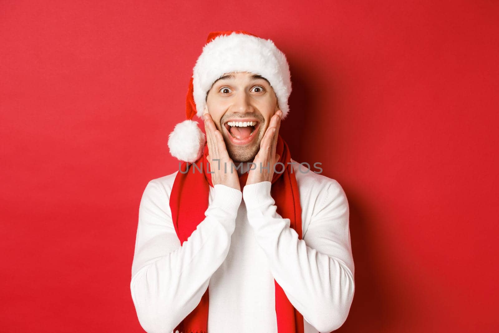 Concept of christmas, winter holidays and celebration. Close-up of surprised and happy man in santa hat and scarf, looking at something amazing, standing over red background.