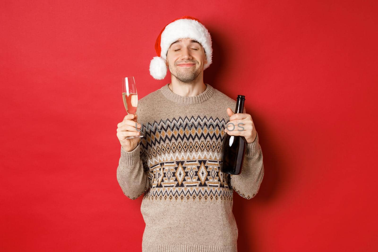 Concept of winter holidays, christmas and celebration. Image of pleased smiling man in santa hat and sweater, drinking on new year, holding bottle of champagne and filled glass, red background.