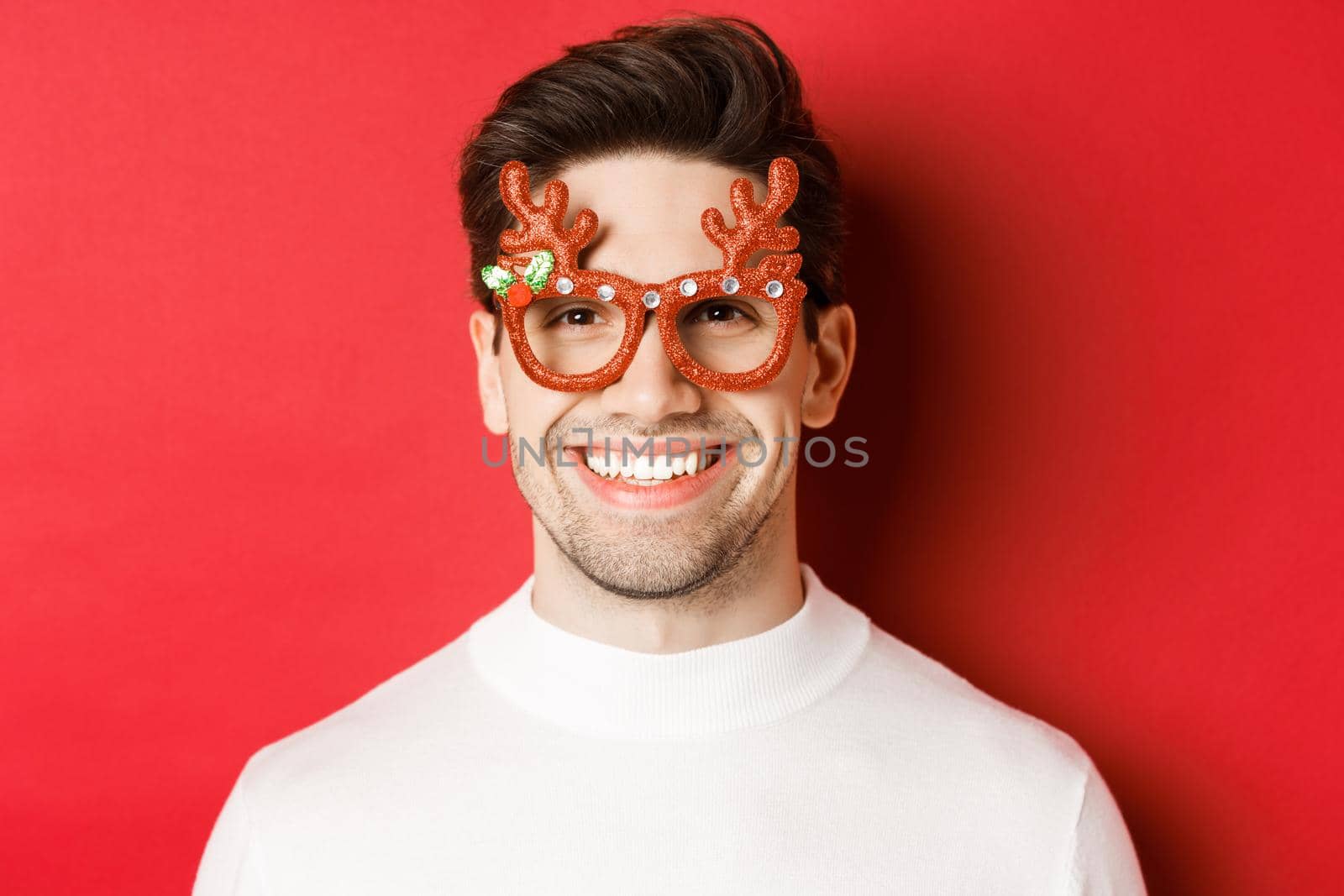 Concept of winter holidays, christmas and celebration. Close-up of attractive brunette guy in party glasses, smiling and looking happy, standing against red background by Benzoix
