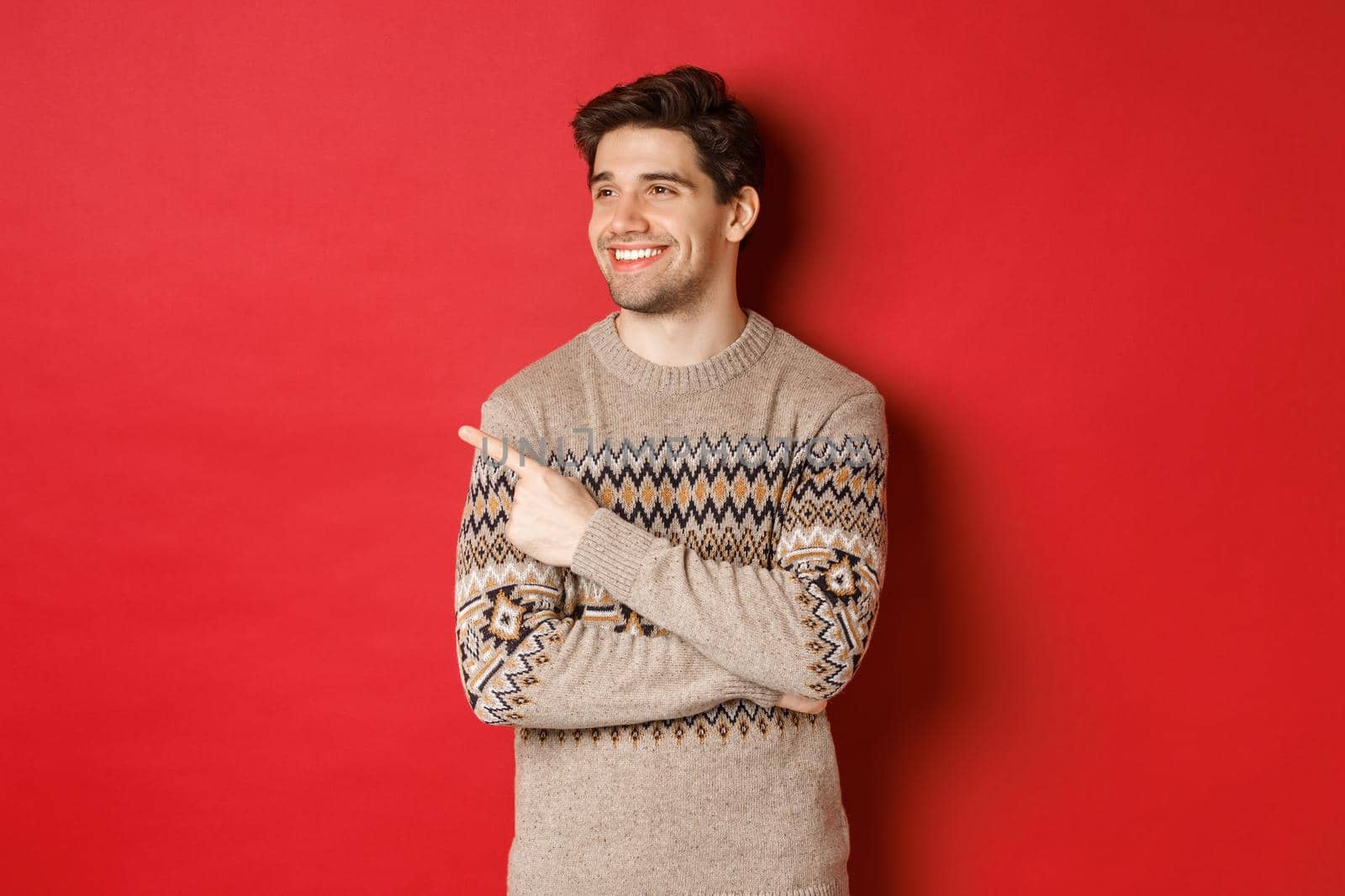 Portrait of attractive caucasian man celebrating new year, winter holidays, pointing finger and looking left with pleased smile, showing christmas advertisement, standing over red background by Benzoix