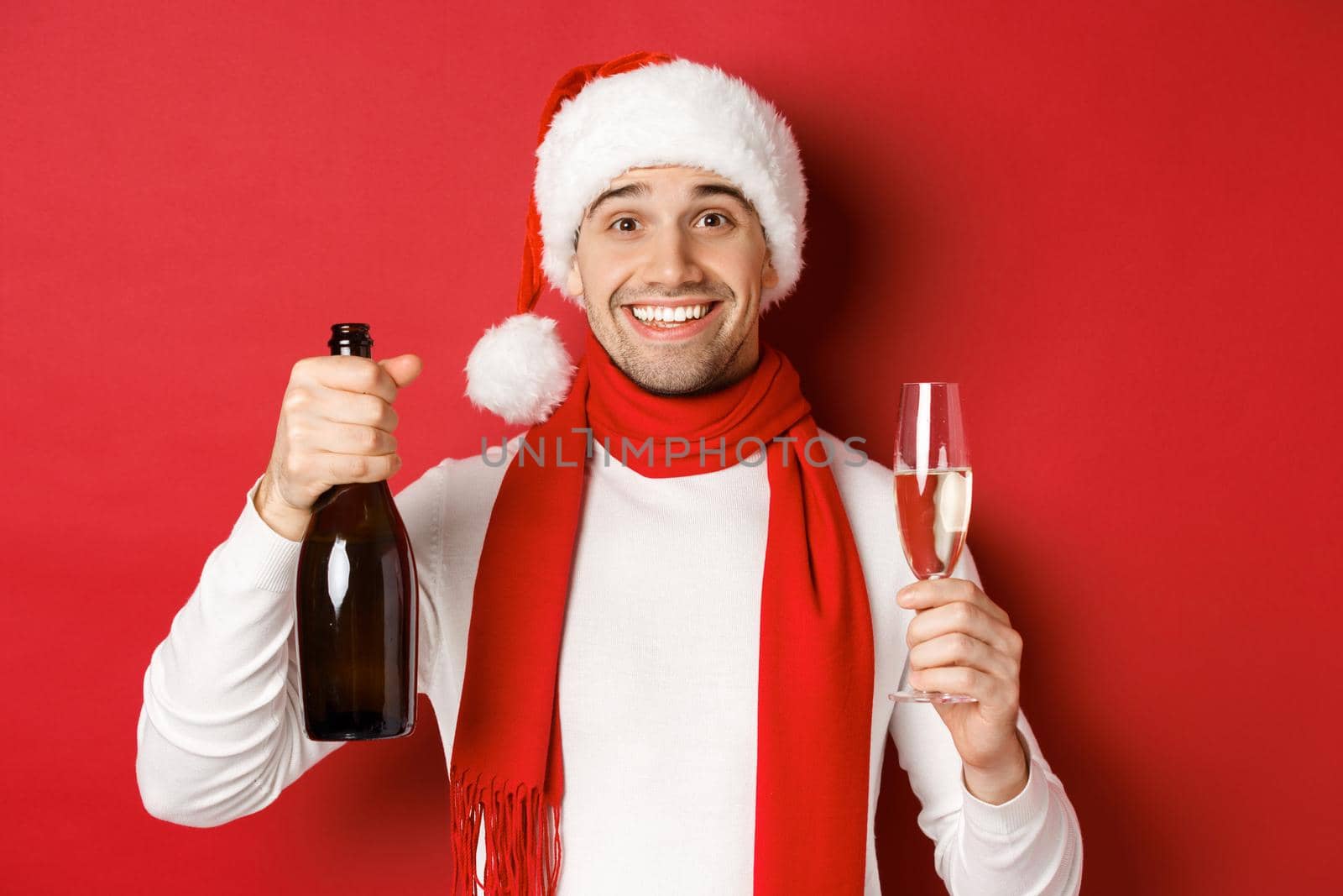 Concept of winter holidays, christmas and lifestyle. Close-up of cheerful handsome man, holding champagne bottle and glass, making toast for new year and celebrating, red background.