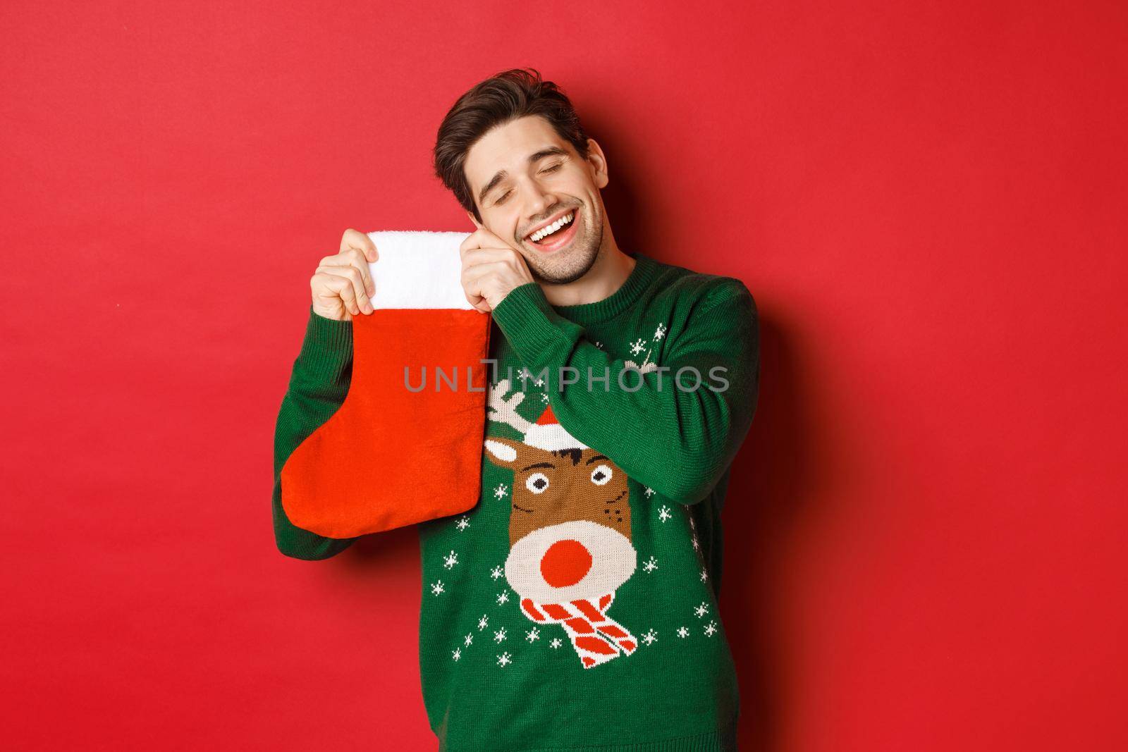 Portrait of happy attractive man in green sweater, looking delighted to receive gifts in christmas stocking, standing over red background.