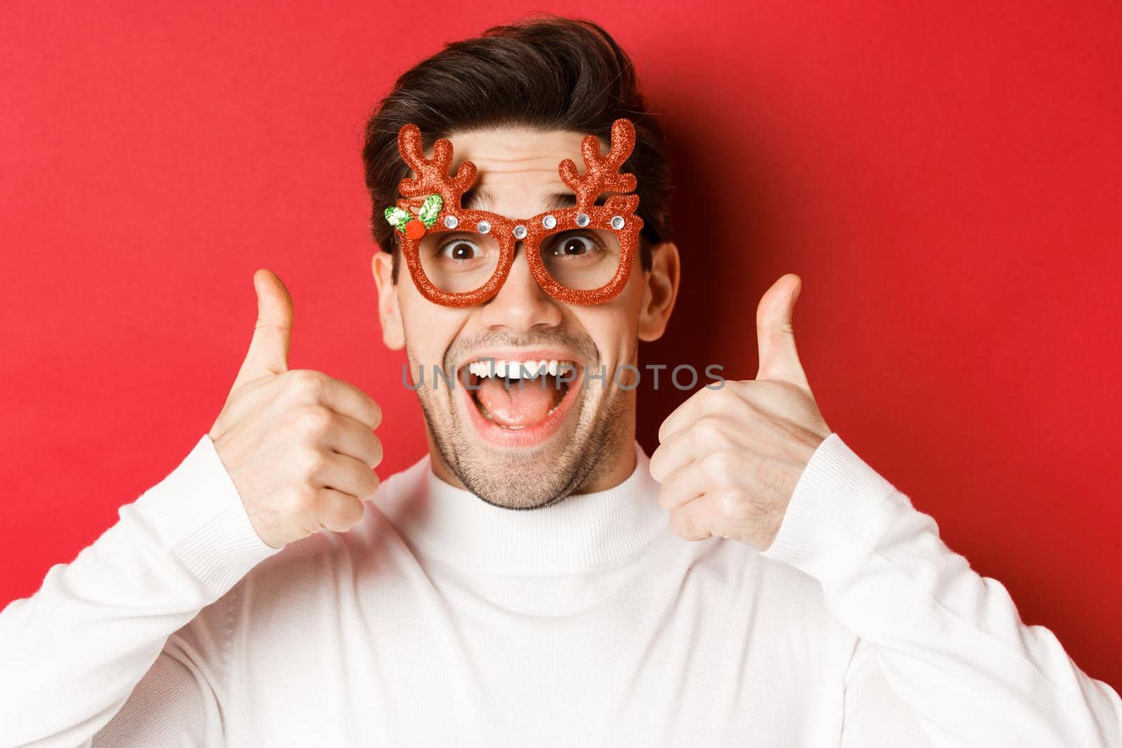 Concept of winter holidays, christmas and celebration. Close-up of excited handsome man in party glasses, smiling and showing thumbs-up in approval, standing over red background by Benzoix