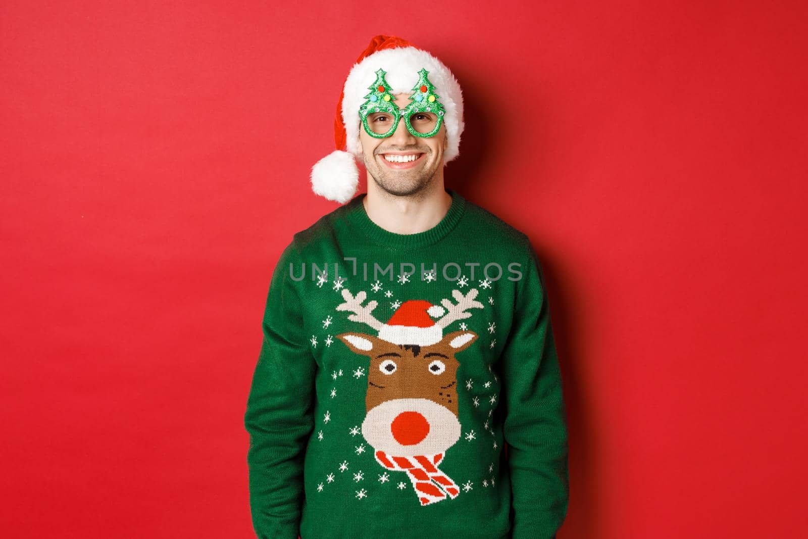 Portrait of handsome man in green christmas sweater, santa hat and party glasses, smiling happy, standing over red background.