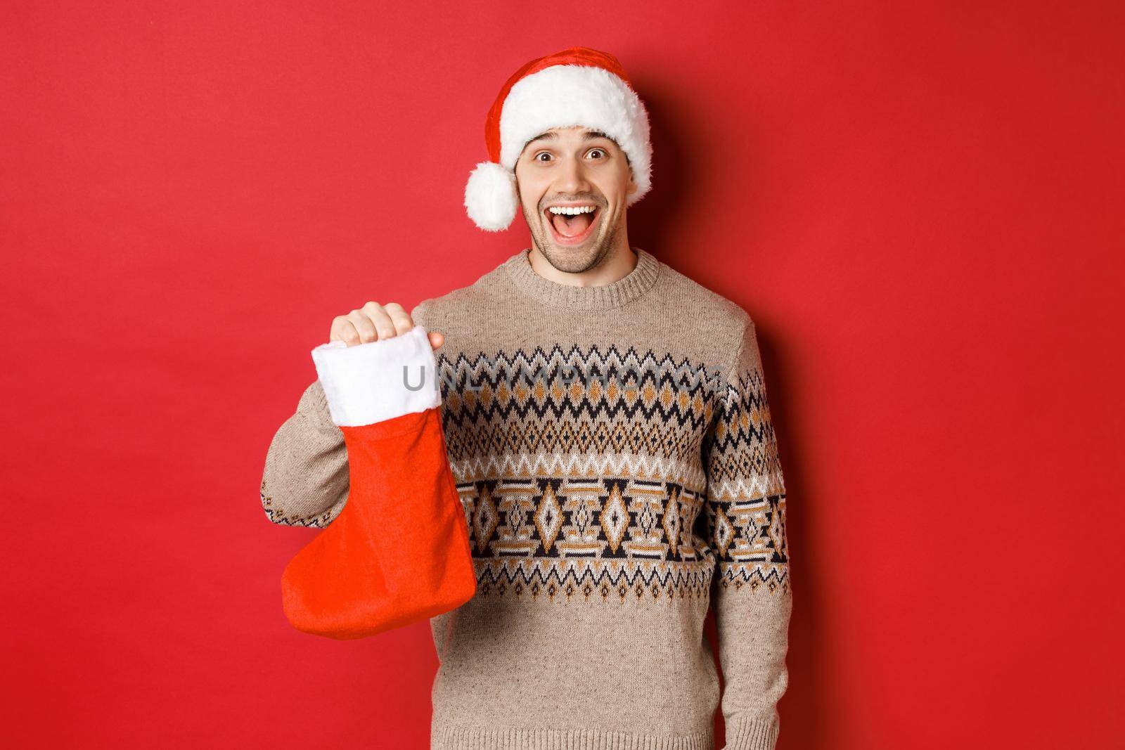 Concept of winter holidays, new year and celebration. Cheerful and surprised adult man receiving candies on Saint Nicholas day in red stocking, standing amazed over red background by Benzoix