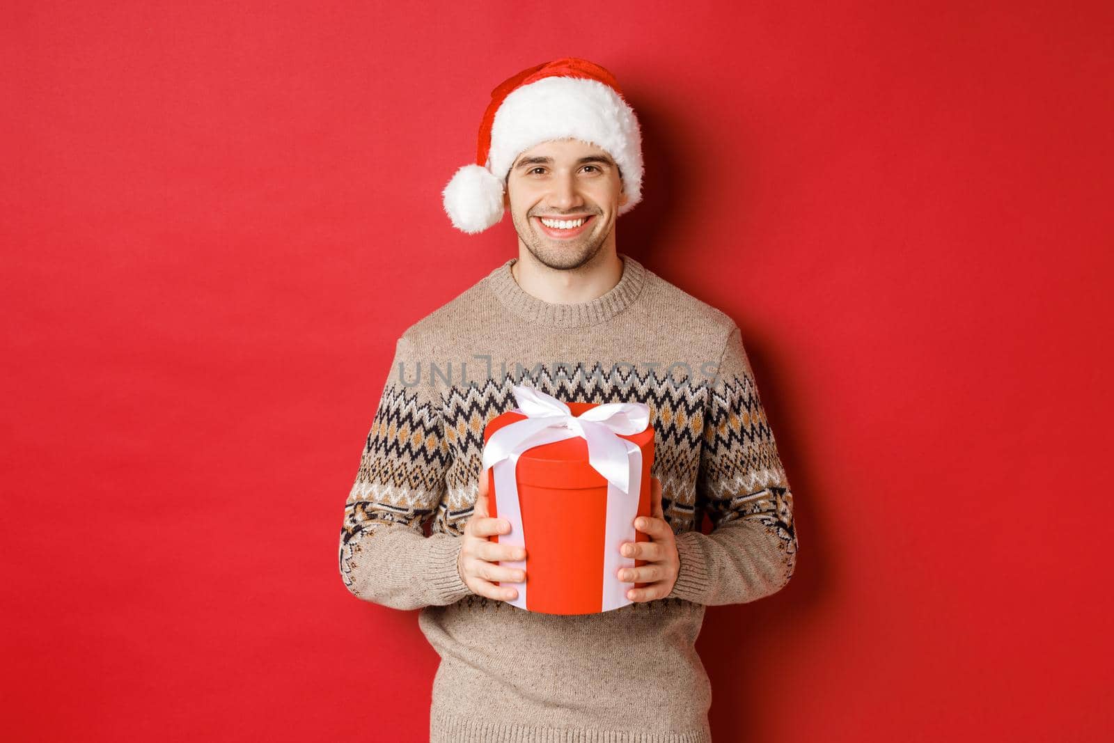 Portrait of handsome man holding a present, wishing happy holiday, standing in santa hat and christmas sweater against red background.