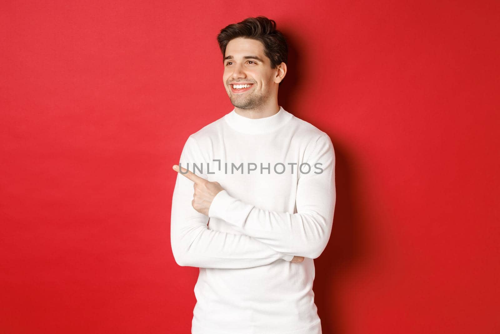 Concept of winter holidays, christmas and lifestyle. Handsome adult man in white sweater, looking and pointing left with pleased smile, showing new year advertisement, red background.