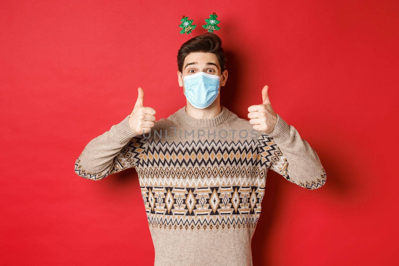 Concept of new year, covid-19 and social distancing. Cheerful caucasian man in sweater and medical mask, celebrating christmas during pandemic, showing thumbs-up, red background.