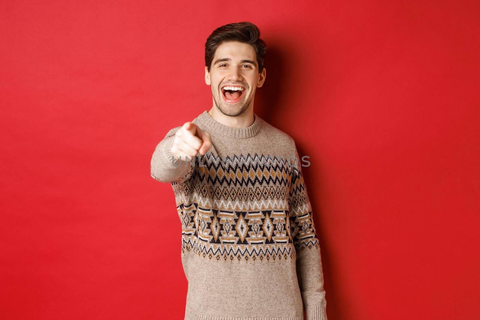 Concept of christmas celebration, winter and lifestyle. Happy attractive man in xmas sweater, wishing happy holidays and pointing at camera, standing over red background.