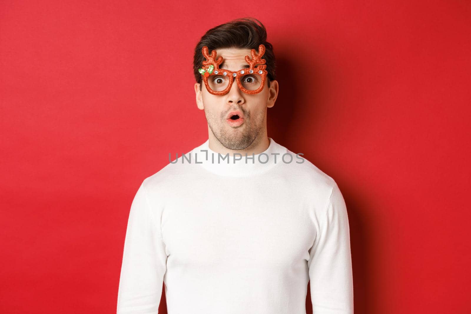 Close-up of surprised and impressed man in white sweater, party glasses, saying wow and looking amazed at camera, standing over red background.