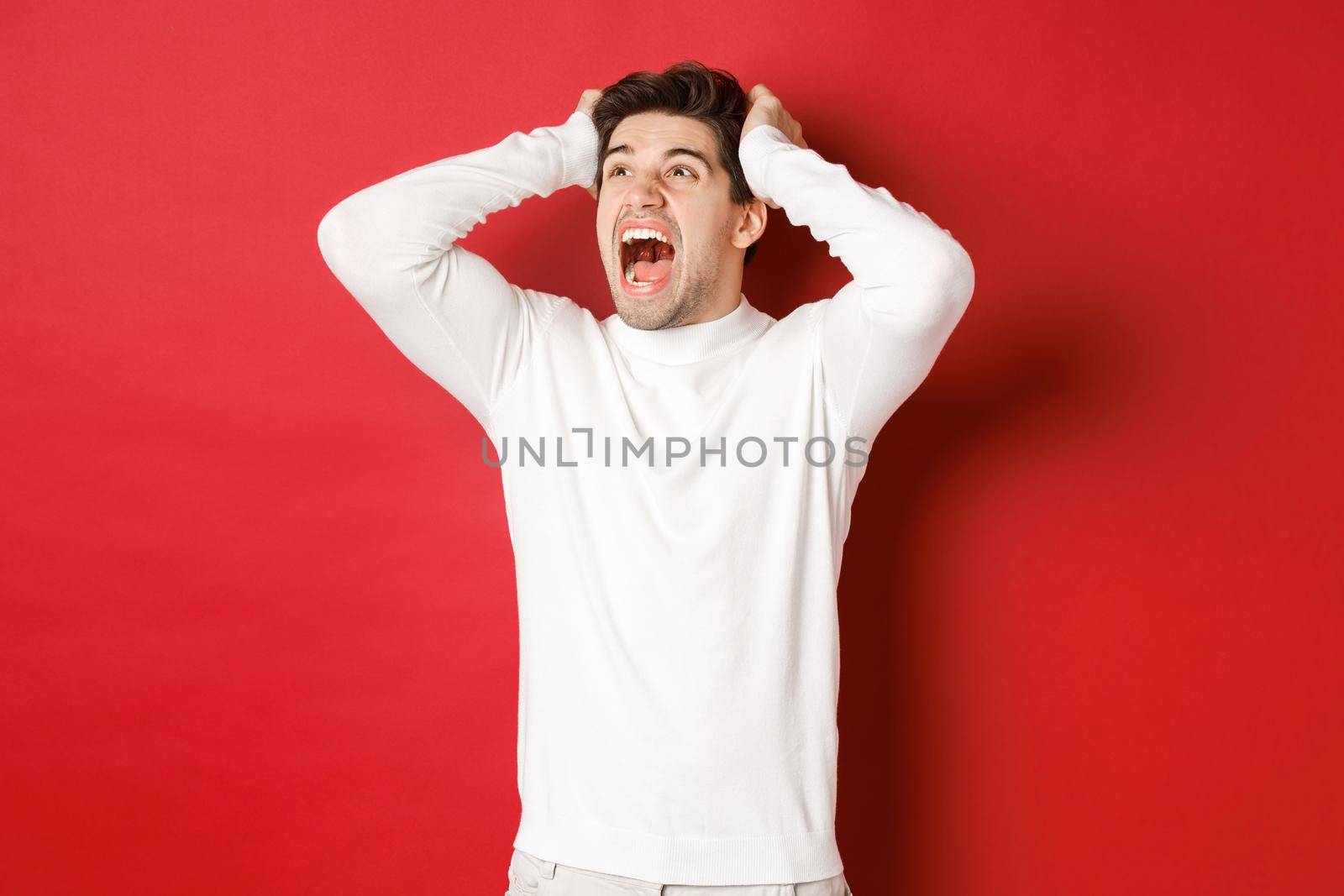Portrait of frustrated man in white sweater, screaming in panic and looking at upper left corner, have terrible problem, standing over red background.