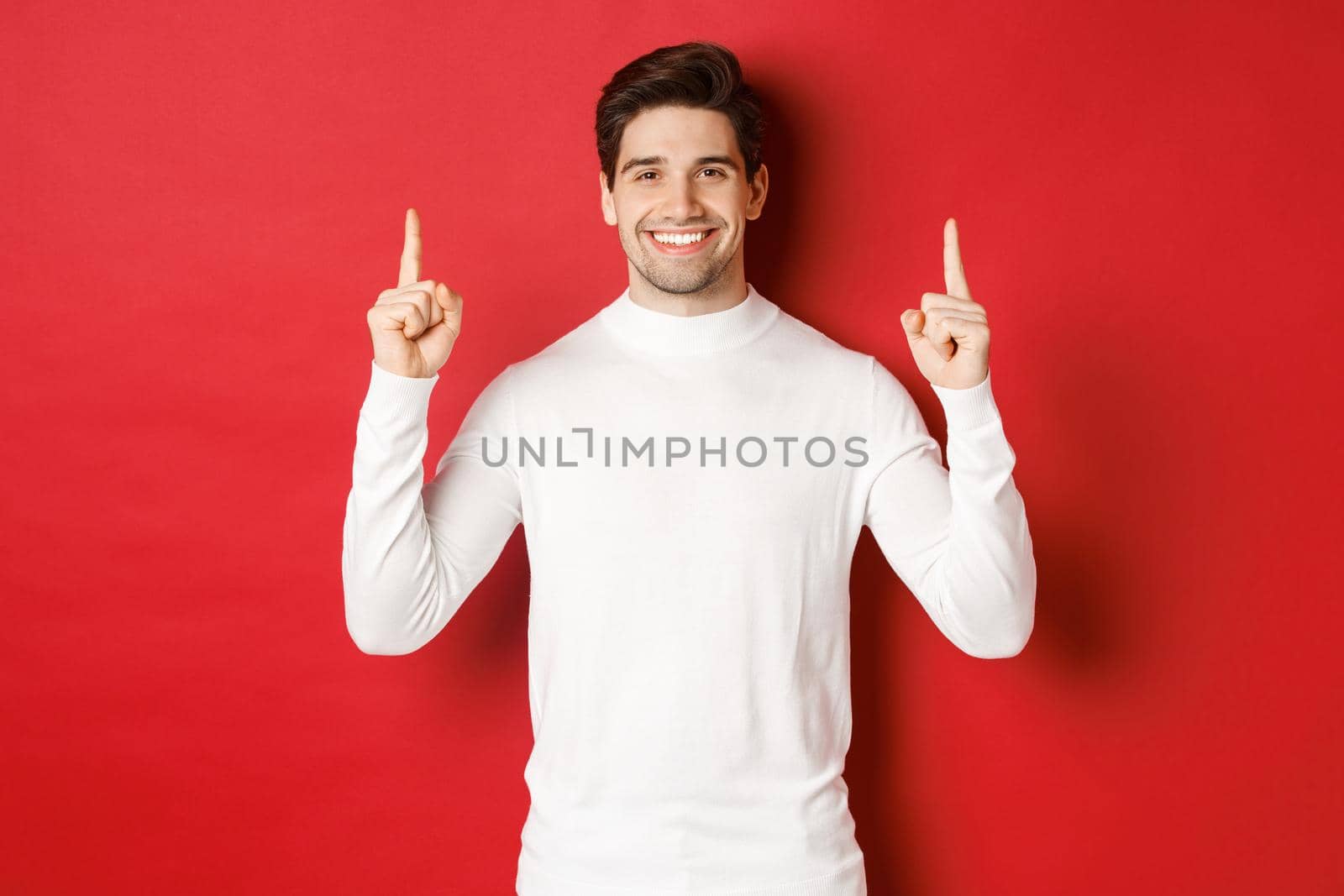 Concept of winter holidays. Good-looking young man with bristle, wearing white sweater, showing christmas advertisement on copy space, pointing fingers up and smiling, red background.