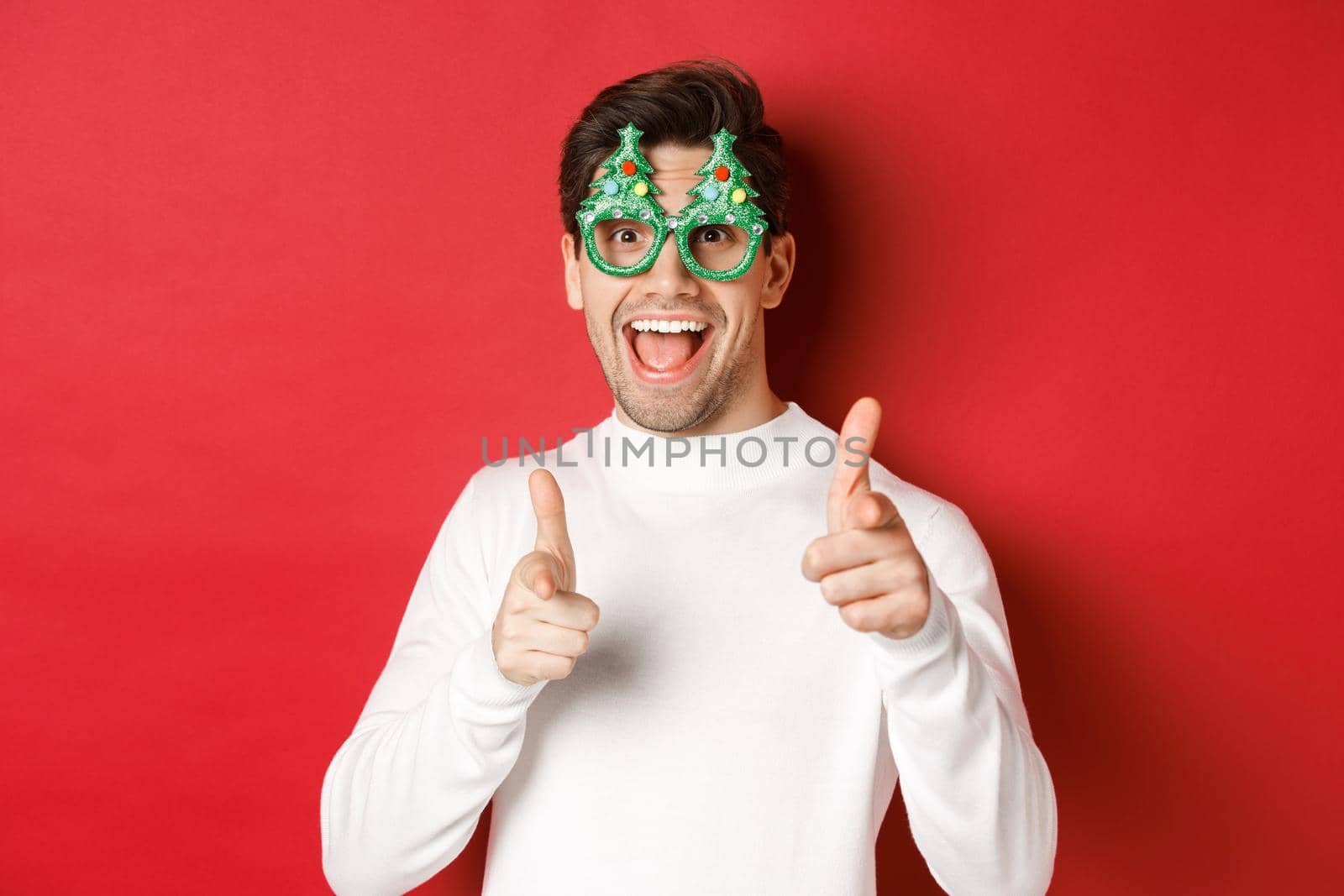 Joyful caucasian guy in party glasses and white sweater, smiling and pointing fingers at camera, wishing merry christmas and happy new year, standing over red background.