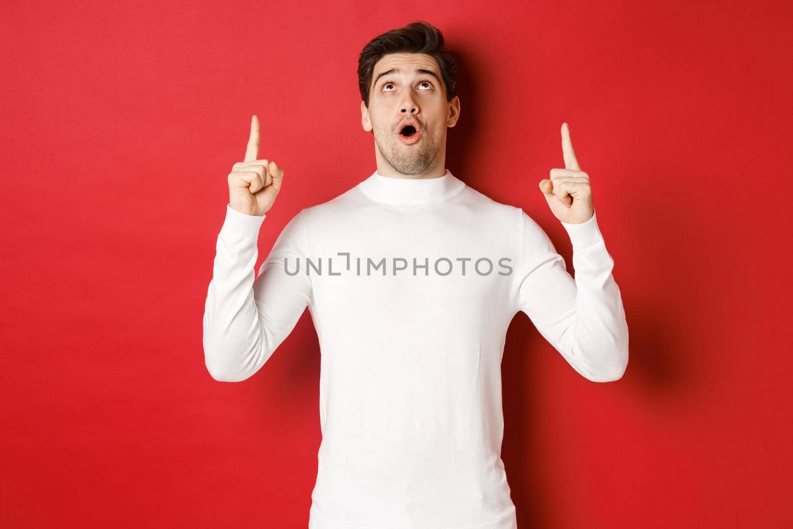 Concept of winter holidays. Portrait of handsome surprised man in white sweater, reacting to new year promo offer, looking and pointing fingers up, standing impressed against red background.