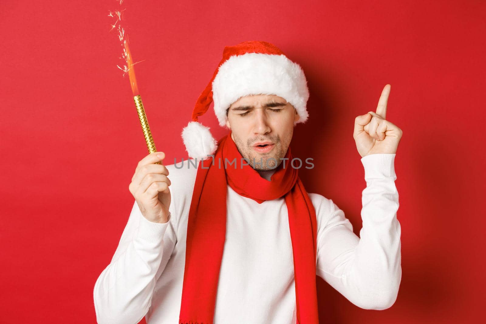 Concept of christmas, winter holidays and celebration. Attractive guy enjoying new year party, dancing with sparkler, wearing santa hat and scarf, standing over red background.