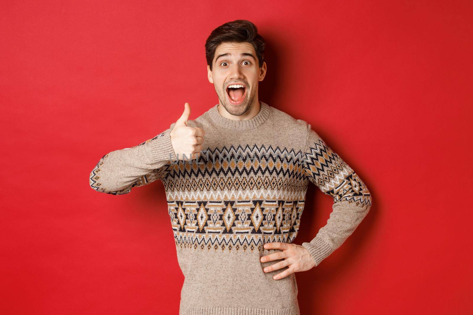 Portrait of excited, attractive man in christmas sweater, showing thumbs-up and looking amazed, recommending good thing, advertising christmas related product.