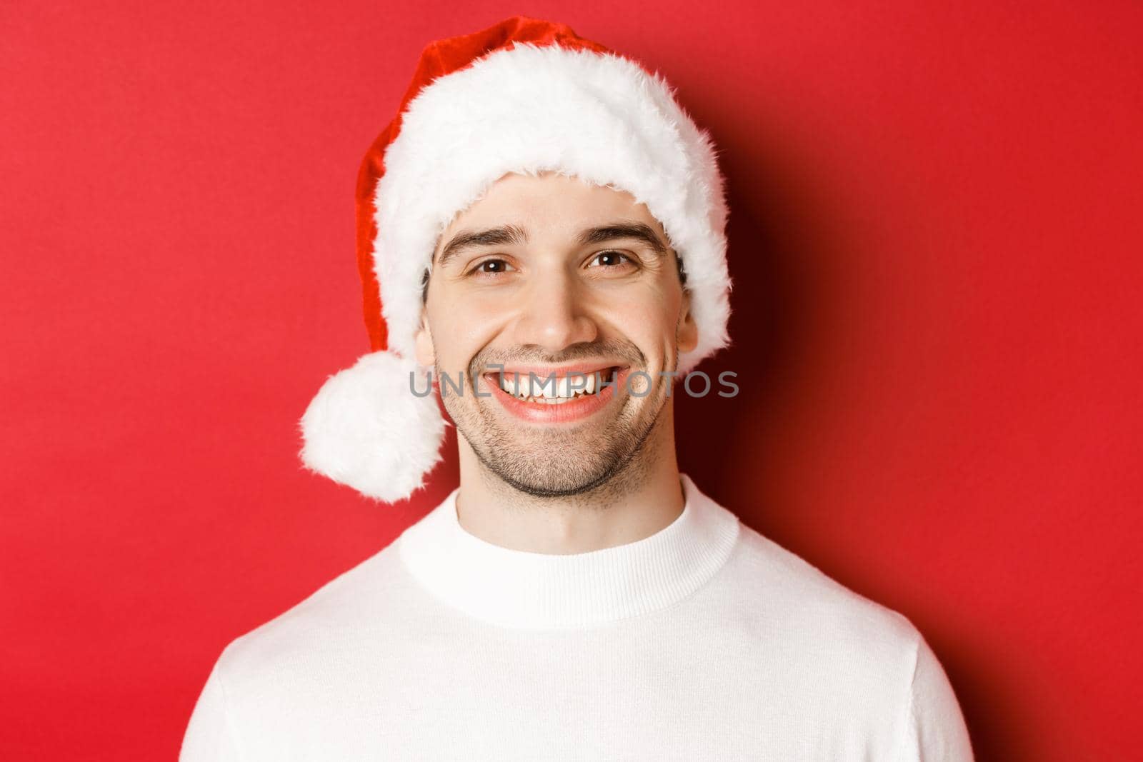 Close-up of attractive smiling man in white sweater and santa hat, looking happy, enjoying winter holidays, standing against red background.