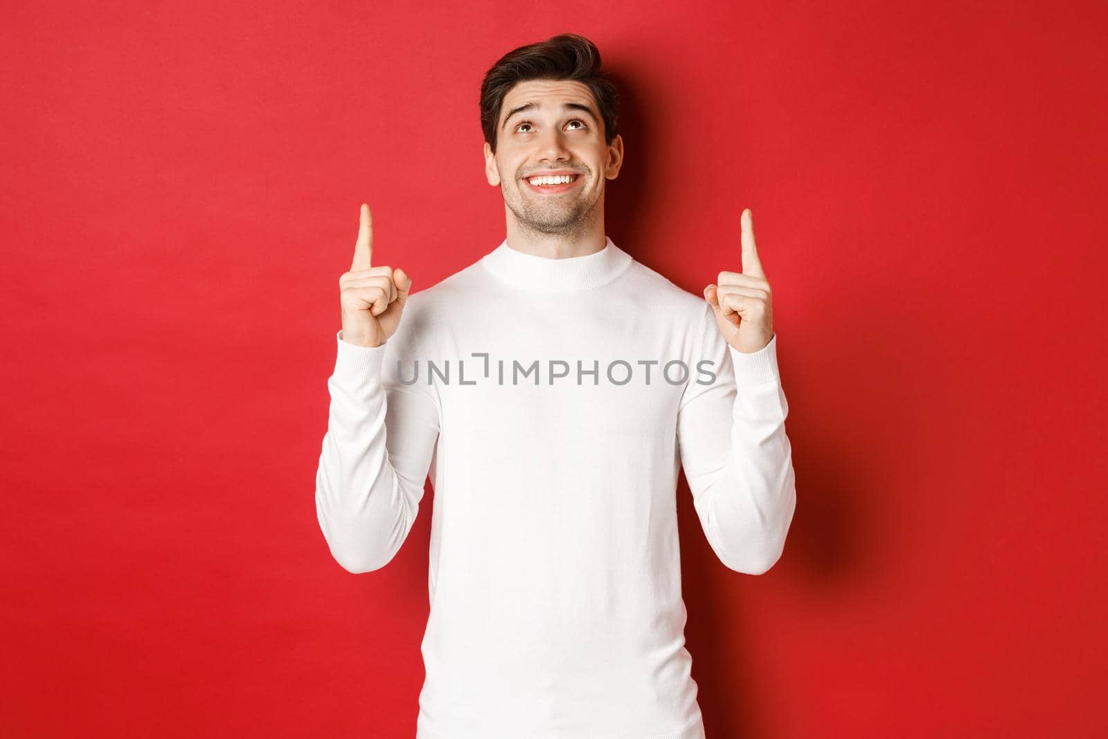Concept of winter holidays. Image of happy attractive man in white sweater, smiling while pointing and looking up at copy space on red background.