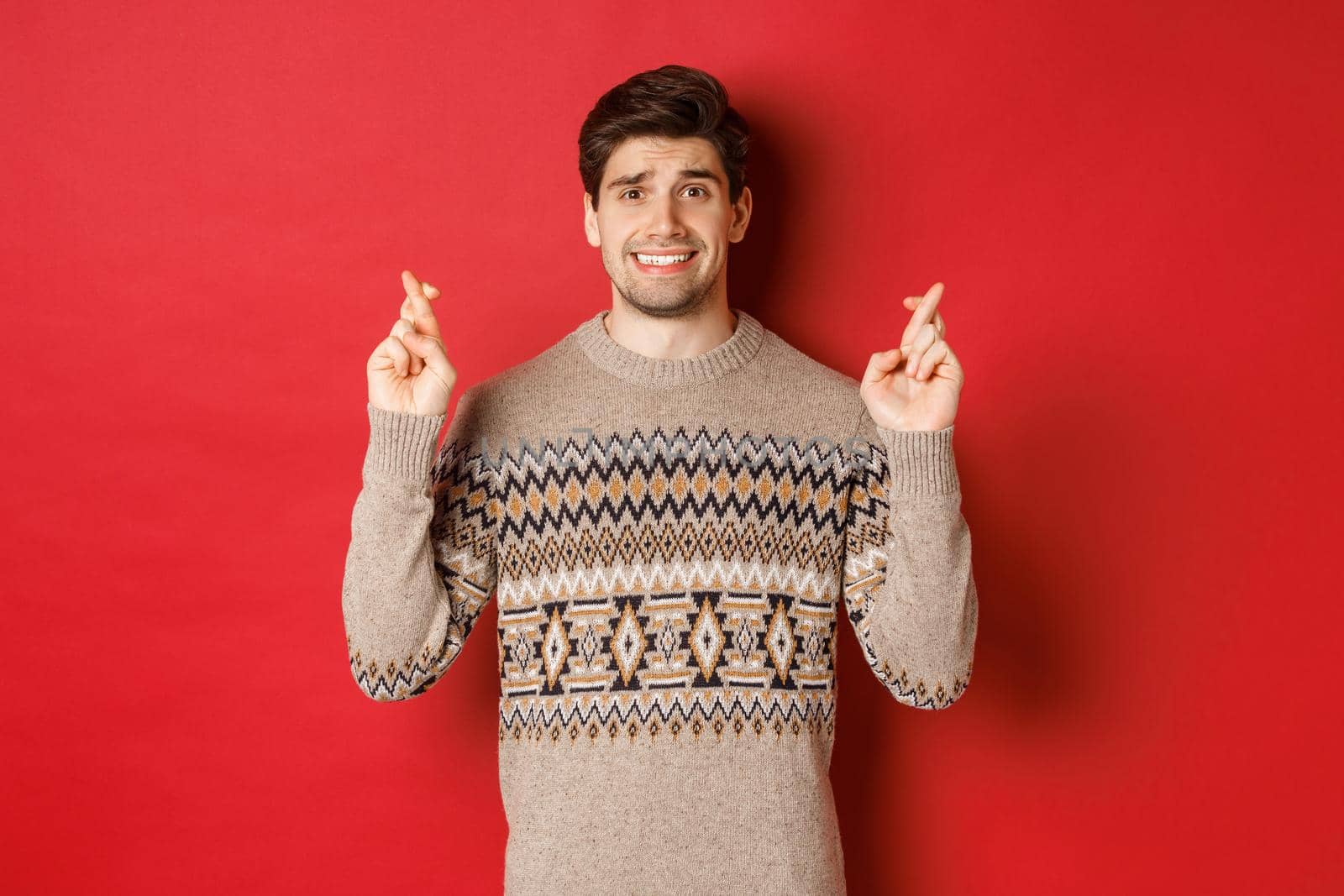 Image of hopeful and worried man in christmas sweater waiting for something, cross fingers for good luck and making wish, nervous about new year gift, red background.