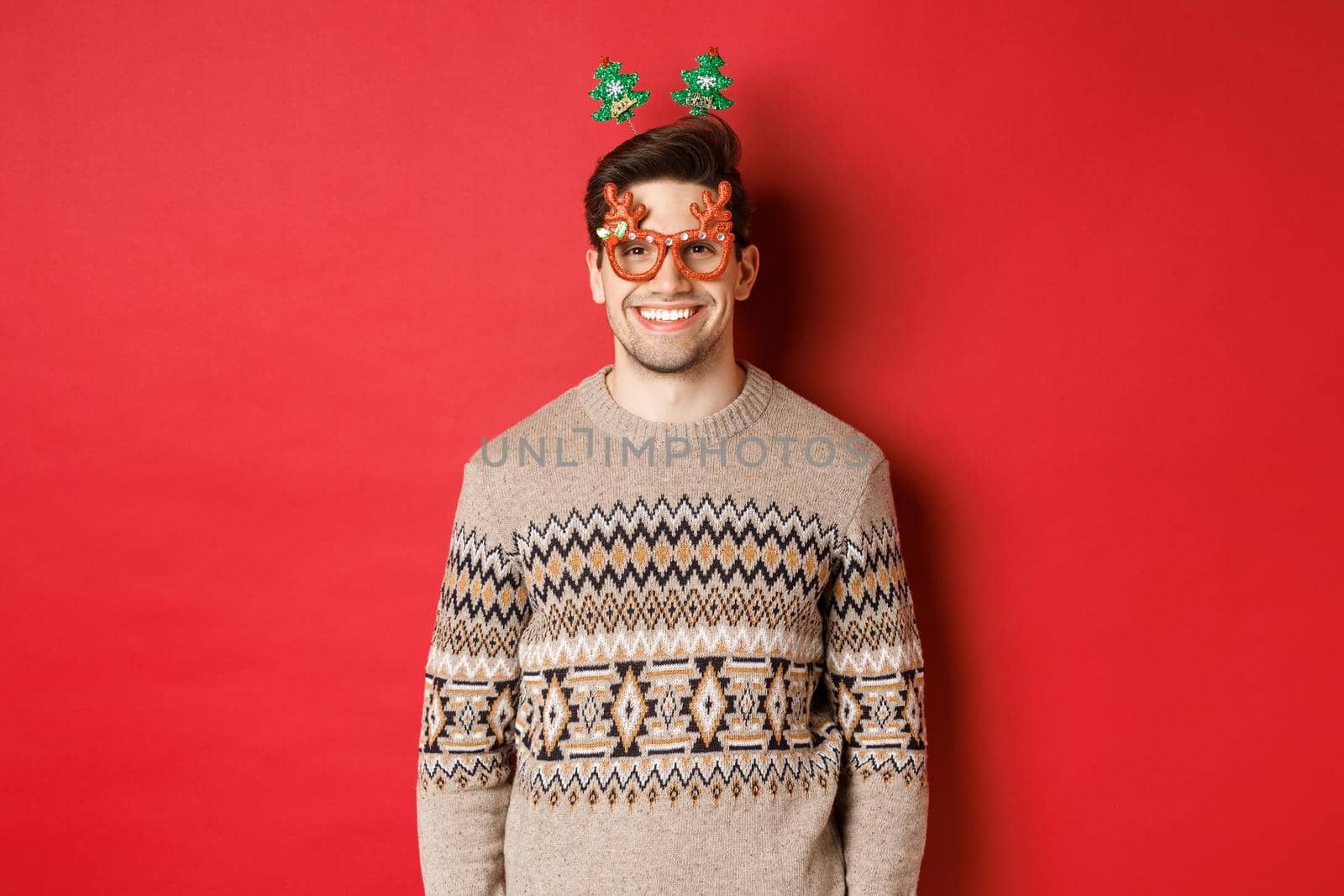 Portrait of handsome bearded guy in party glasses and winter sweater, smiling pleased, celebrating new year, wishing merry christmas, standing over red background by Benzoix
