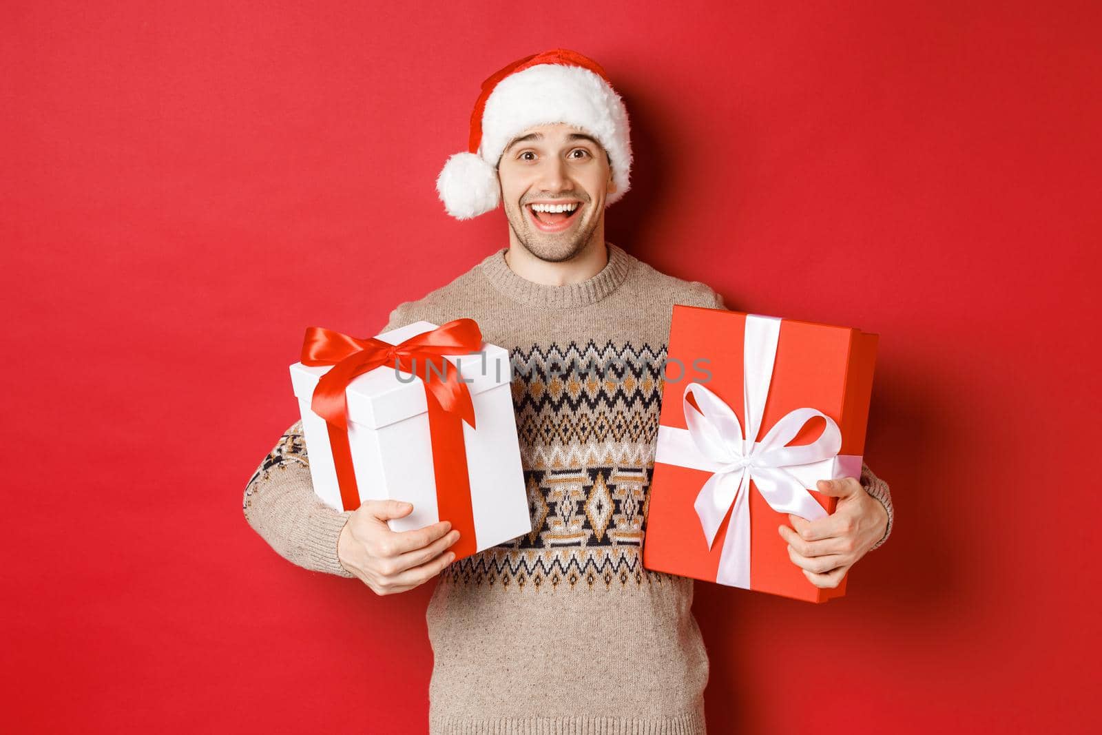 Image of cheerful attractive guy holding christmas presents, standing in santa hat and winter sweater, smiling happy, standing over red background.