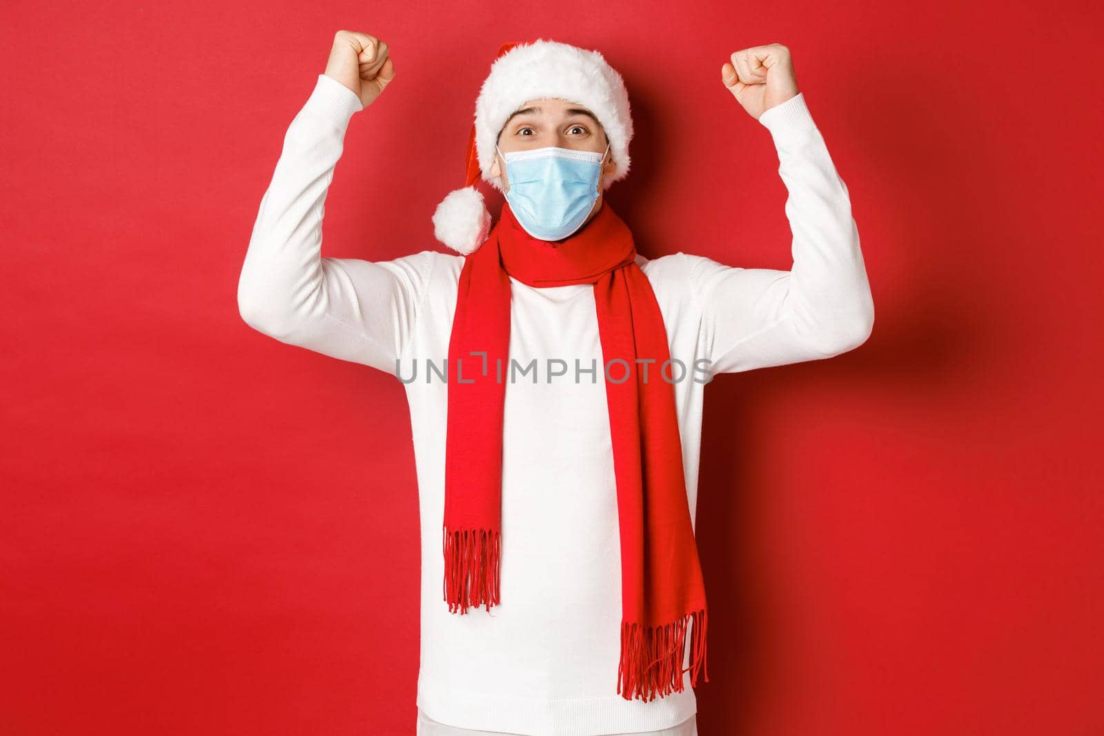 Concept of covid-19, christmas and holidays during pandemic. Portrait of happy man in santa hat and medical mask, rejoicing and celebrating new year, standing over red background.