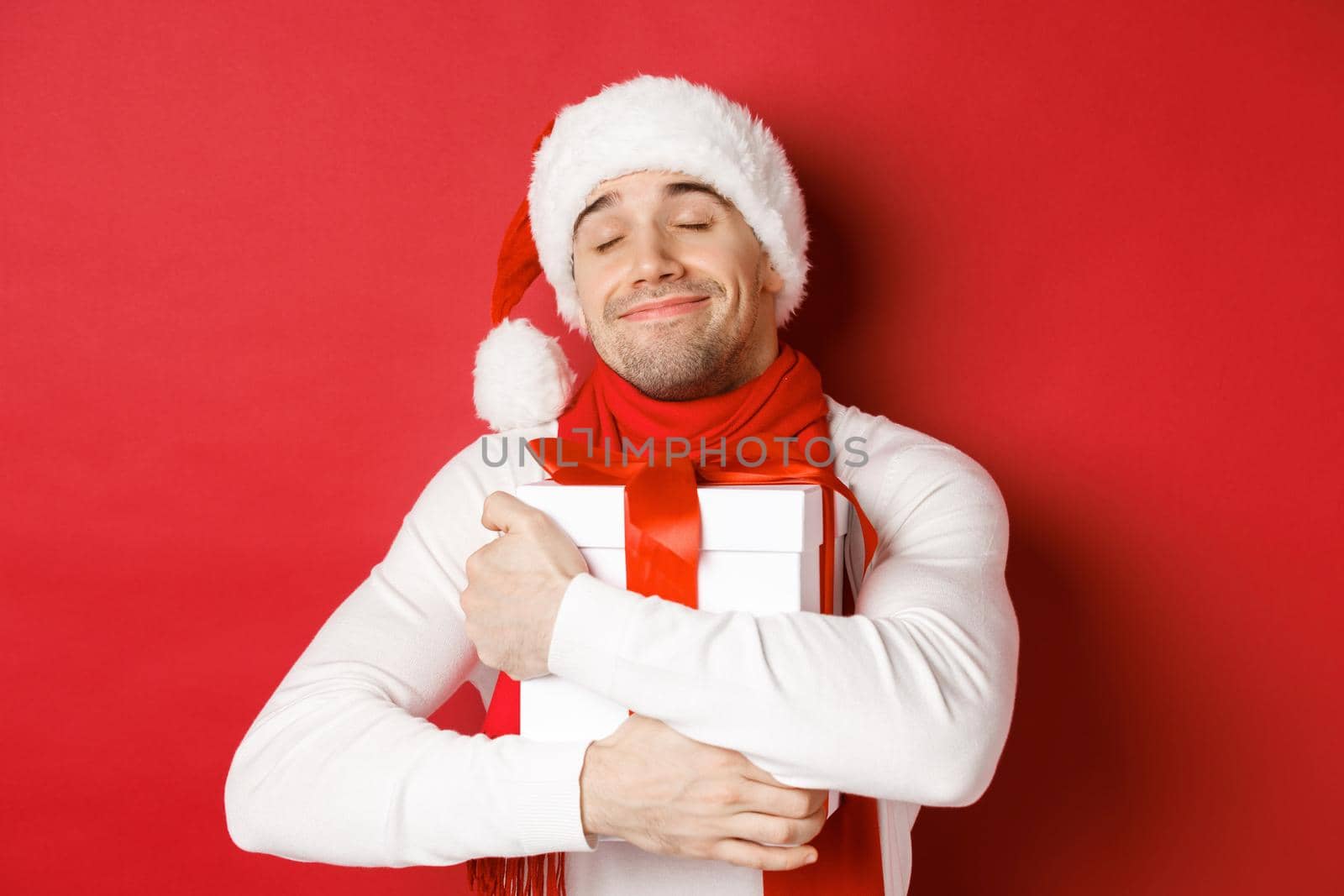 Concept of winter holidays, christmas and lifestyle. Image of lovely guy in santa hat and scarf, hugging his new year present and smiling flattered, standing over red background. Copy space