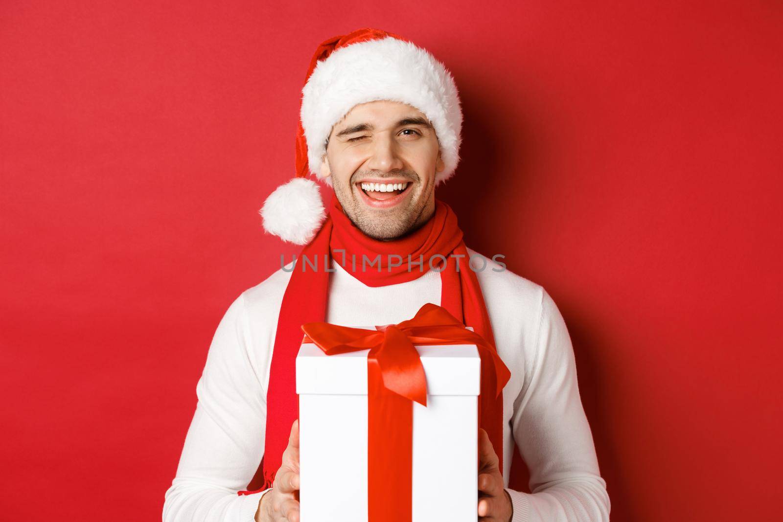 Concept of winter holidays, christmas and lifestyle. Handsome cheeky man in santa hat and scarf, holding present and smiling, winking at camera, standing over red background.